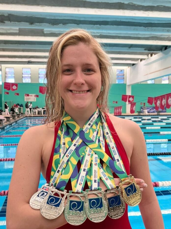 #descricaodaimagem #pratodosverem Foto de Mariana com várias medalhas. Ao fundo vemos a piscina. Mariana é loira e está sorrindo.