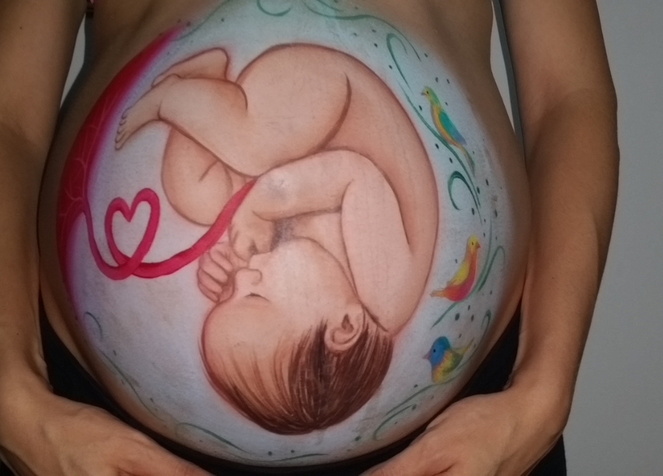 #descricaodaimagem #pratodosverem foto de uma barriga de gestante pintada com a imagem de um bebê. O cordão umbilical é cor de rosa e forma um coração.