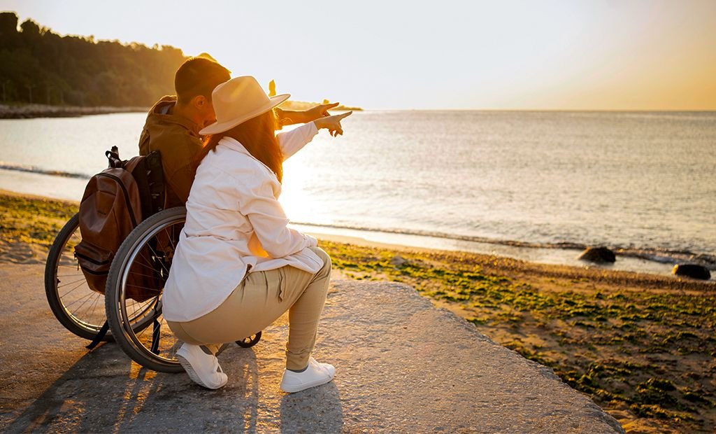 #descricaodaimagem #pratodosverem foto de um casal na praia. Eles estão lado a lado e apontam para o horizonte. O homem é cadeirante e a mulher está abaixada, ao seu lado.