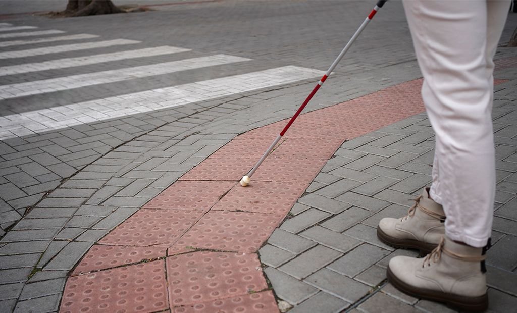 #descricaodaimagem #pratodosverem foto de uma pessoa cega, usando uma bengala, está caminhando em uma calçada com acessibilidade para pessoas cegas.
