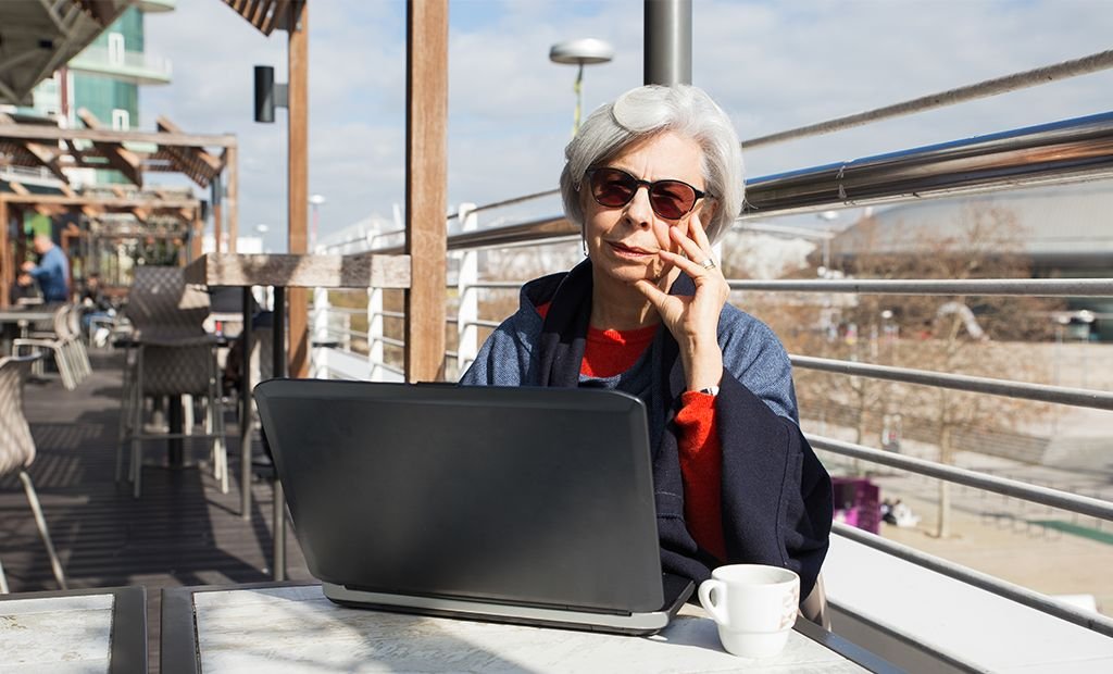 #descricaodaimagem #pratodosverem foto de uma mulher idosa. Ela está em um café e usa um notebook que está aberto sobre a mesa.