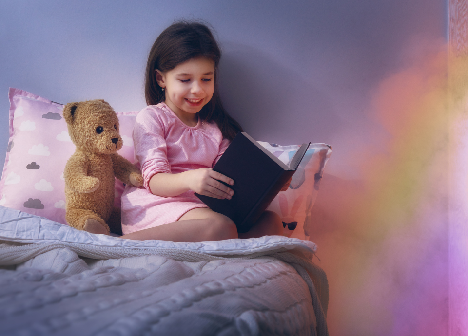 #descricaodaimagem #pratodosverem foto de uma menina lendo. Ela está na cama e, ao seu lado, está um urso de pelúcia. A foto tem um efeito em tons de lilás rosa e amarelo.