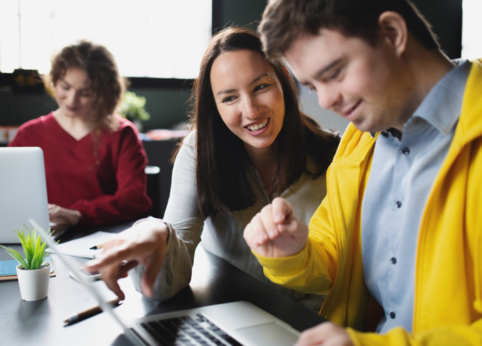 #descricaodaimagem #pratodosverem foto de um ambiente corporativo com três pessoas. Em primeiro plano, uma mulher e um homem estão olhando algo num notebook. Ele tem síndrome de Down. Ao fundo está uma outra mulher, usando um notebook.