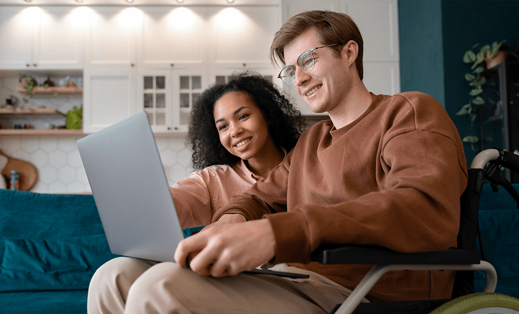#descricaodaimagem #pratodosverem foto de um homem e uma mulher sentados lado a lado. Eles estão vendo a tela de um notebook. O rapaz é cadeirante.