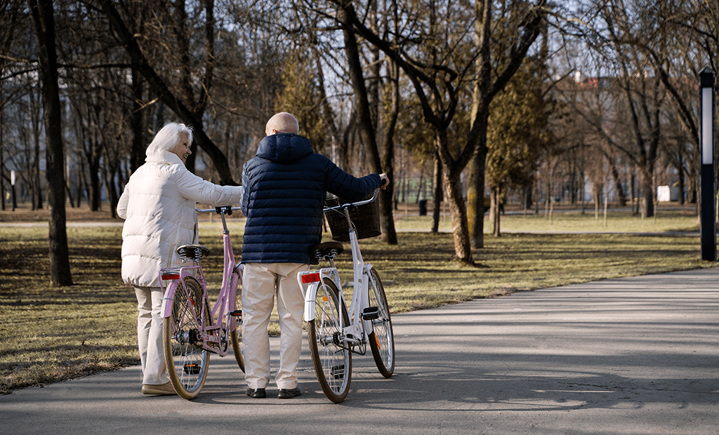 #descricaodaimagem #pratodosverem foto de um casal de idosos caminhando em um parque e empurrando uma bicicleta. O casal usa roupas de frio e caminha entre árvores.