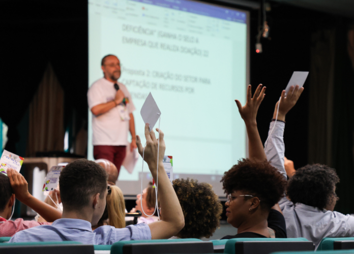 #descricaodaimagem #pratodosverem foto da conferência, no momento de votação das propostas. Em 1º plano estão os participantes, alguns deles com as mãos levantadas, segurando seus crachás. Em 2º plano está o presidente do conselho, no palco, segurando o microfone. Atrás dele está um telão.