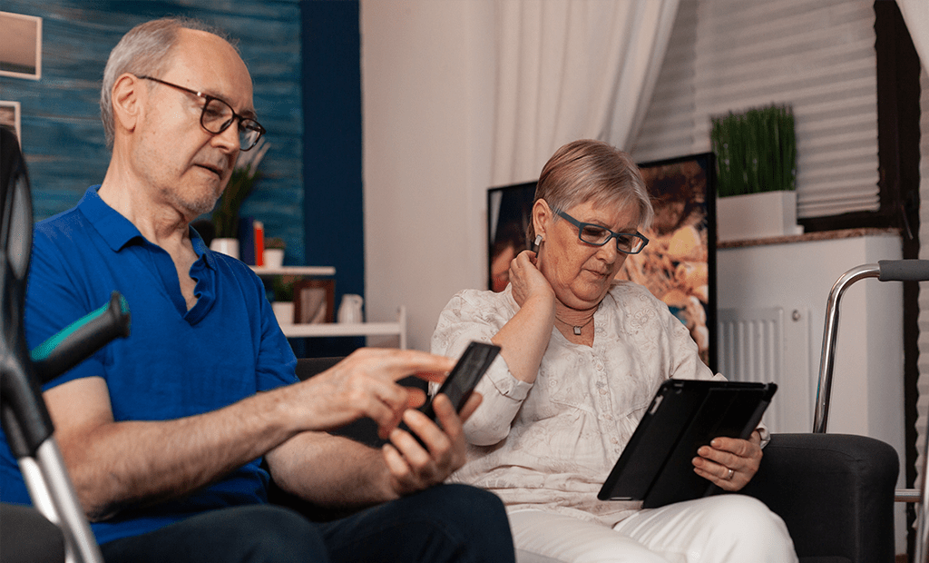 #descricaodaimagem #pratodosverem foto de um casal de idosos sentado na sala mexendo em aparelhos eletrônicos. O homem está mexendo em um celular e a mulher mexendo em um tablet.