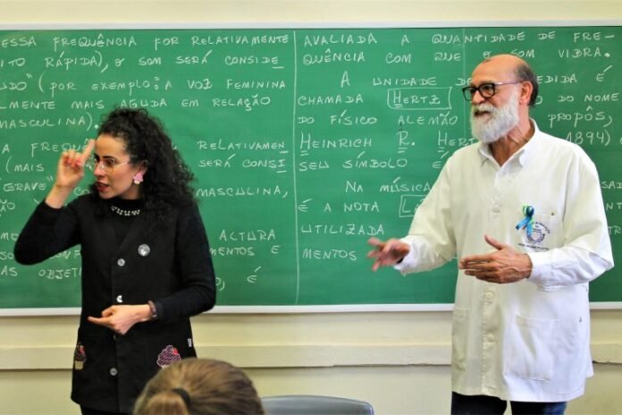 #descricaodaimagem #pratodosverem foto de uma sala de aula com um professor e uma intérprete de Libras, durante uma explicação. Atrás deles está uma lousa verde com várias coisas escritas com giz branco.