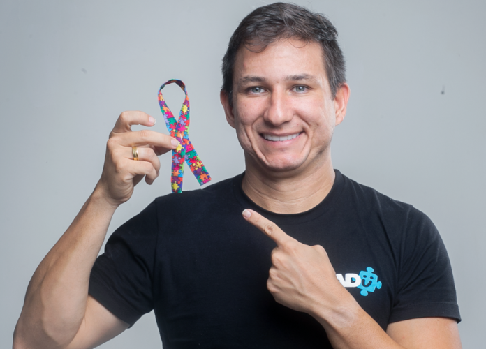 #descricaodaimagem #pratodosverem foto do diretor do Instituto do Autismo, Kadu Lins. Ele é um homem branco, que está sorrindo e segura um laço que simboliza o autismo.
