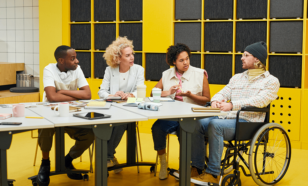 #descricaodaimagem #pratodosverem foto de quatro pessoas conversando em uma sala de reunião. São dois homens e duas mulheres. Um dos homens é cadeirante. A sala tem parede amarela com detalhes em preto.