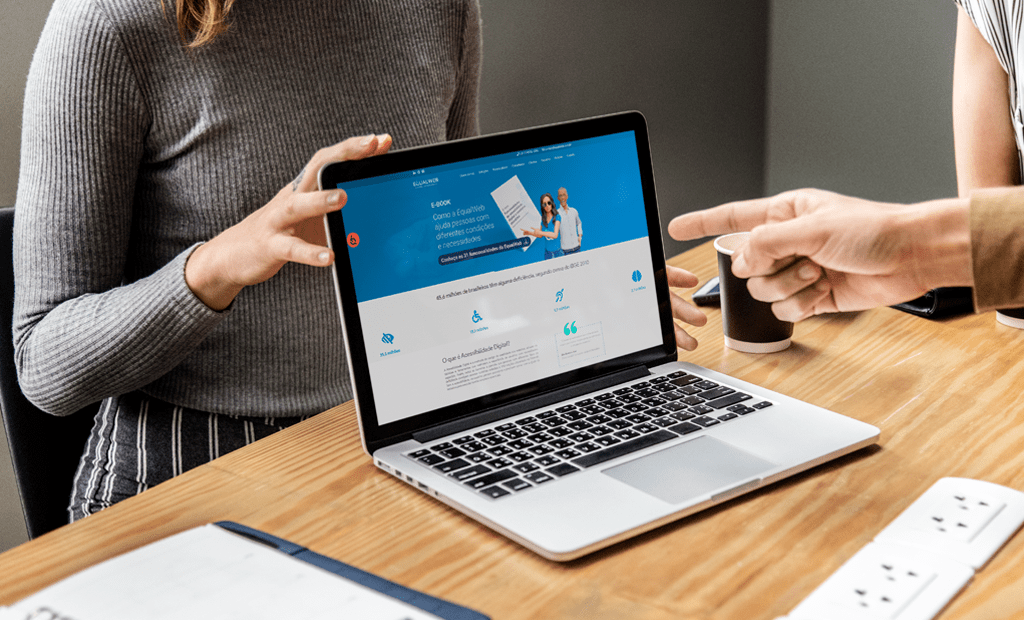 #descricaodaimagem #pratodosverem foto de um notebook aberto em cima de uma mesa com a página do site da EqualWeb aberta. Em volta da mesa aparecem algumas mãos de pessoas apontando para o site.