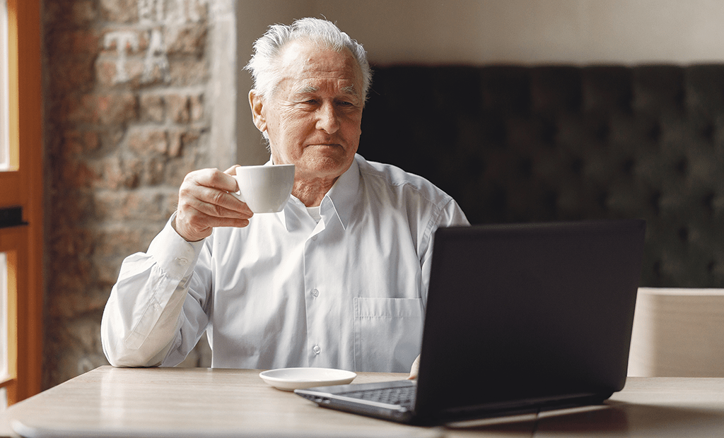 #descriçãodaimagem #pratodosverem foto de um homem branco, de cabelos grisalhos, tomando café em frente a um computador.