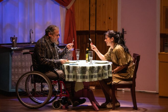 #descriçãodaimagem #pratodosverem foto de uma cena da peça Misery. Na cena os protagonistas estão sentados, jantando, em uma mesa redonda. O homem é cadeirante.