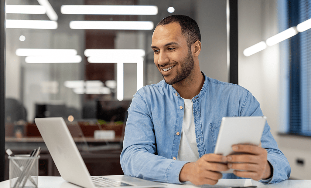 #descriçãodaimagem #pratodosverem foto de um homem em um ambiente corporativo. Ele está segurando um tablet e olha para um notebook. O homem é negro e está vestindo uma camiseta branca com camisa jeans.