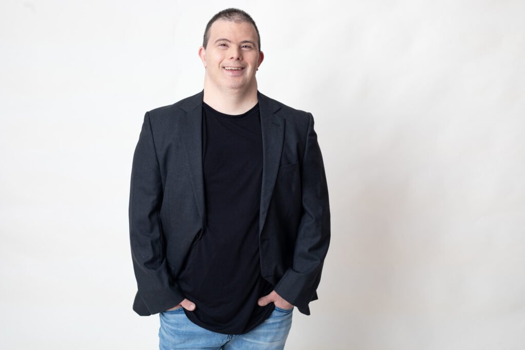 #descriçãodaimagem #pratodosverem foto do Breno Viola. Ele é um homem com síndrome de Down. Breno está vestindo calça jeans, e camiseta e blazer preto.