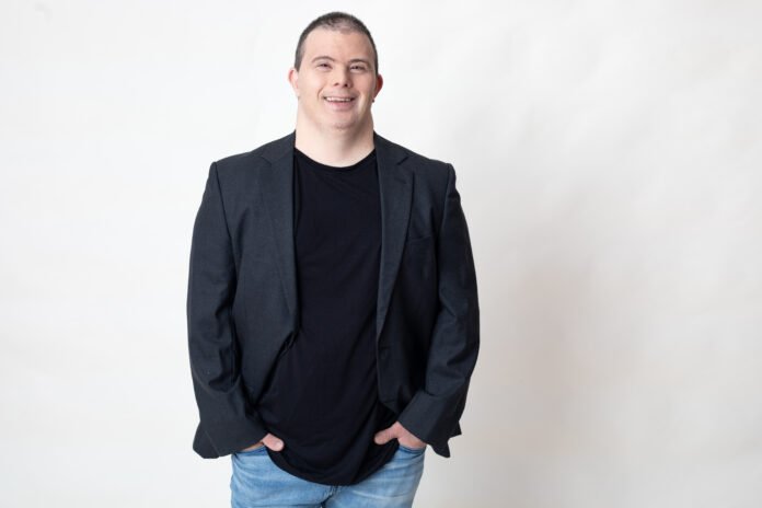 #descriçãodaimagem #pratodosverem foto do Breno Viola. Ele é um homem com síndrome de Down. Breno está vestindo calça jeans, e camiseta e blazer preto.