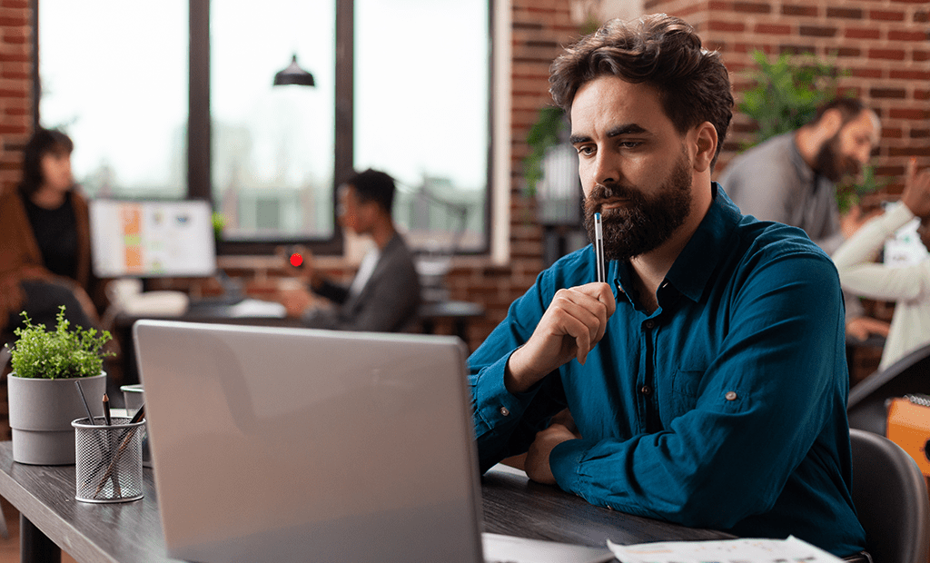 #descriçãodaimagem #pratodosverem foto de um homem em um ambiente corporativo. Ele está usando um notebook. O homem é branco, tem barba e está vestindo uma camisa azul.