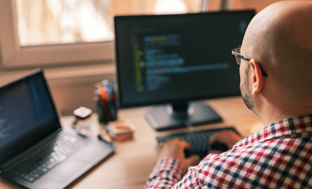 #descriçãodaimagem #pratodosverem foto de um homem branco. Ele é careca, usa óculos e está vestindo uma camisa xadrez. Ele está usando o computador e na imagem aparece duas telas à sua frente.