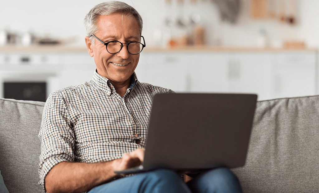 #descriçãodaimagem #pratodosverem foto de um homem idoso com cabelos grisalhos sorrindo e usando óculos, ele está sentado no sofá com um notebook em sua perna. 
