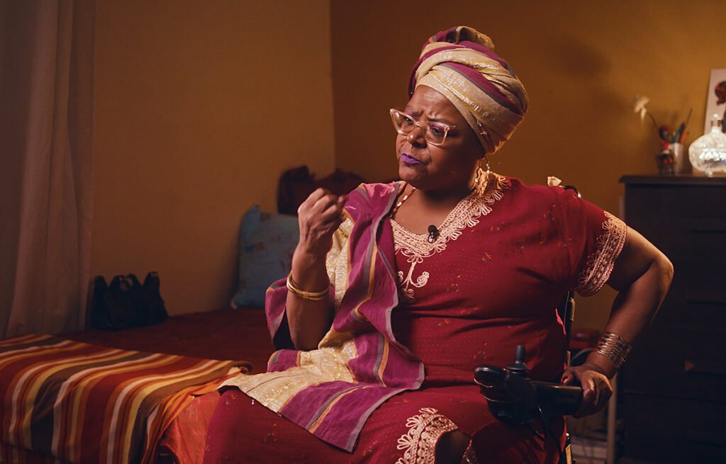 #descriçãodaimagem #pratodosverem foto de Mona Rikumbi. Ela é uma mulher negra, cadeirante, e está vestindo trajes africanos.