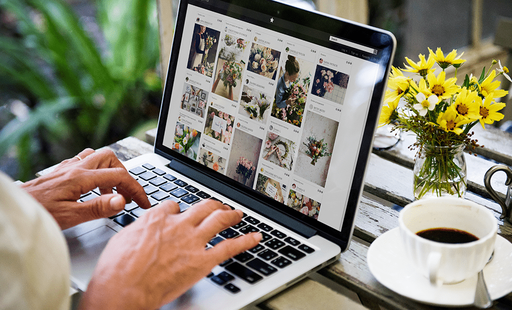 #descriçãodaimagem #pratodosverem foto de uma mão feminina mexendo em um notebook. Ao lado, na mesa, tem uma xícara de café e um vaso com flores amarelas.