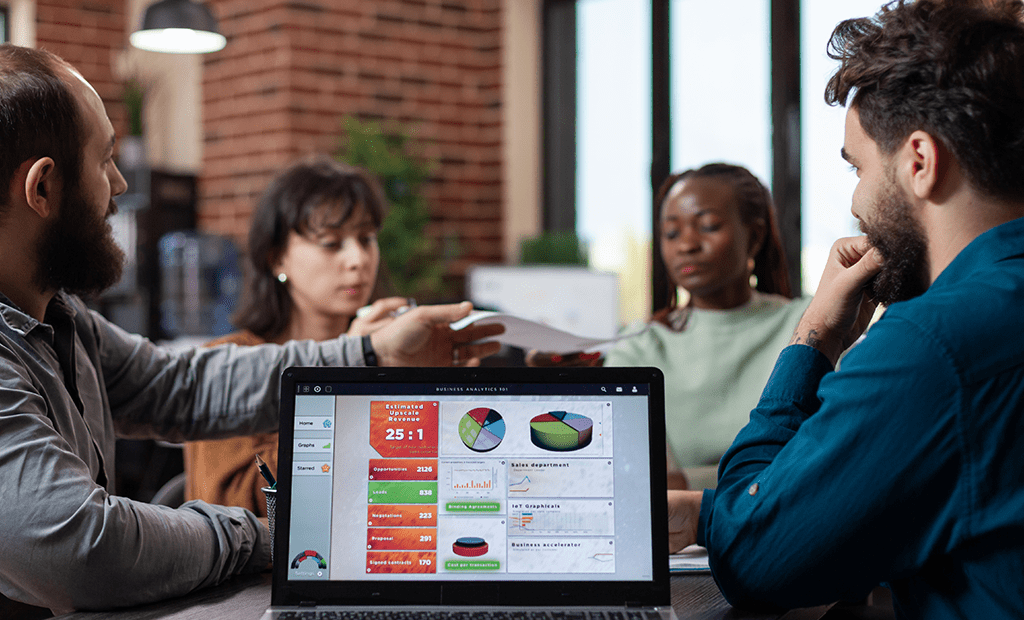 #descriçãodaimagem #pratodosverem foto de quatro pessoas de diferentes etnias em uma reunião de negócios, discutindo gráficos na tela de um laptop.