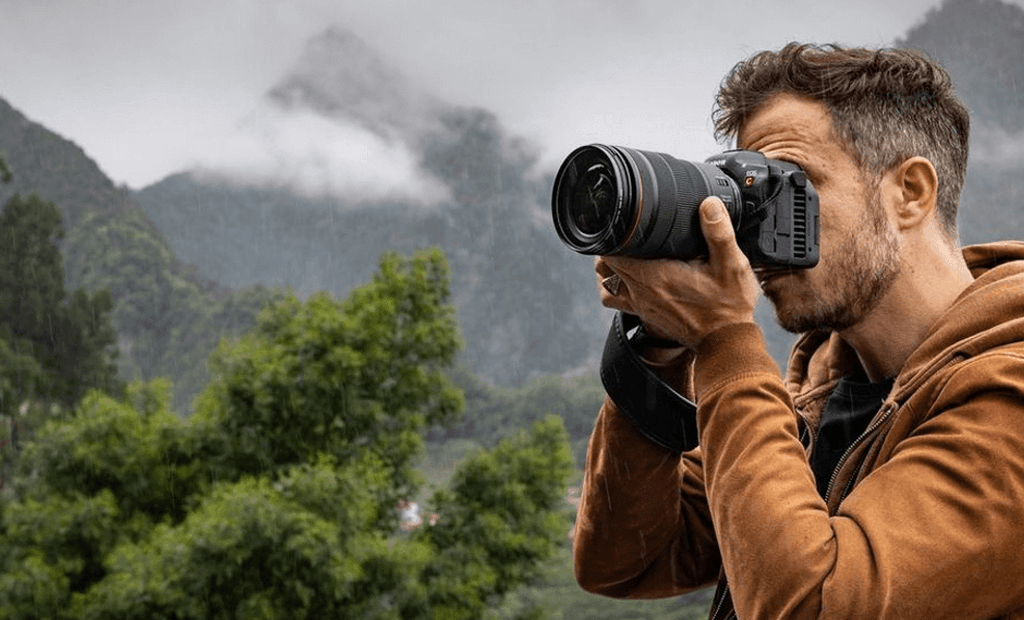 #descriçãodaimagem #pratodosverem foto de um fotógrafo usando a câmera digital Canon EOS R5, na cor preta, para capturar imagens e vídeos de uma bela paisagem em 8K.
