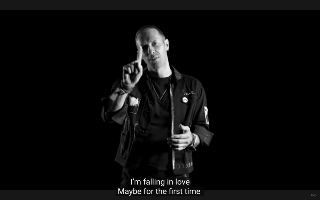 #descriçãodaimagem #pratodosverem imagem em branco e preto do clipe do Coldplay. O vocalista da banda, Chris Martin, sinaliza na língua de sinais.
