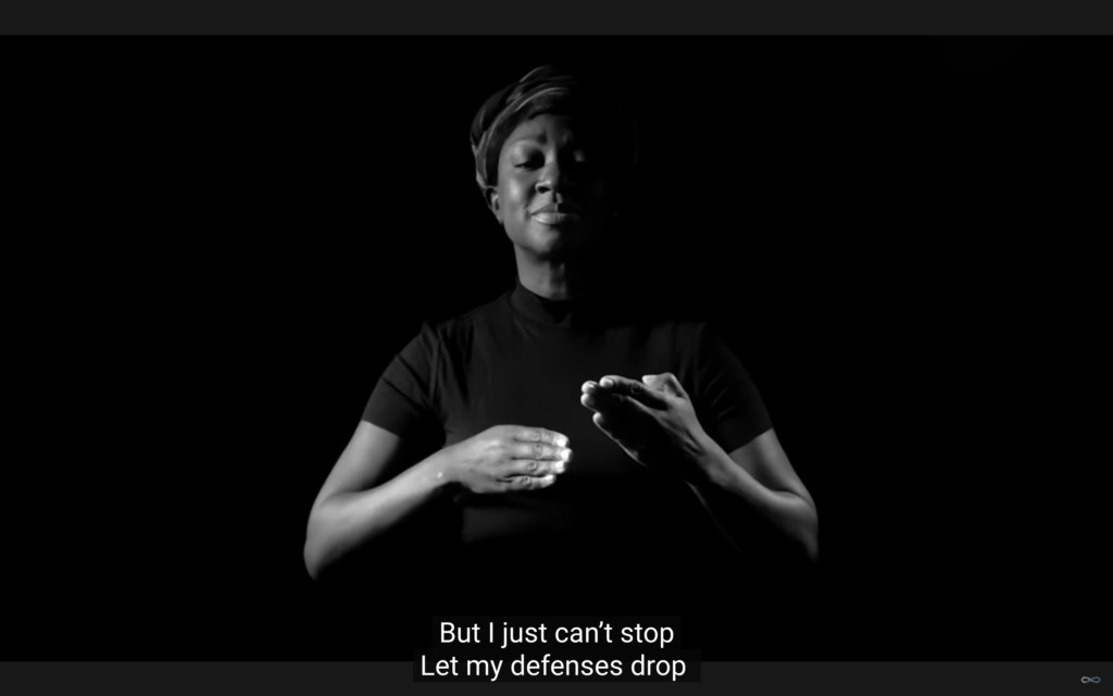 #descriçãodaimagem #pratodosverem imagem em branco e preto do clipe da banda Coldplay. A atriz Natasha Ofili, sinaliza na língua de sinais.