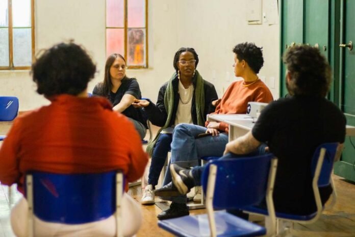 #descriçãodaimagem #pratodosverem registro fotográfico do 3º encontro do curso de acessibilidade cultural. Cinco pessoas sentadas em cadeiras organizadas em círculo. Duas delas estão desfocadas e de costas para a foto. Ao fundo, em foco, três mulheres de frente. Uma mulher branca de cabelos lisos castanhos na altura do ombro. Uma mulher crespa, ao centro, em meio a uma fala. E a 3ª uma mulher parda de cabelos curtos castanhos, estilo moicano. Ela está olhando para a mulher que está ao seu lado.
