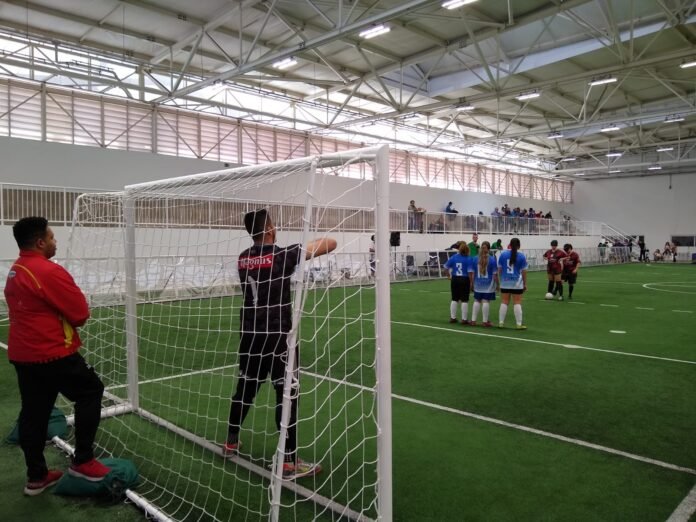 #descriçãodaimagem #pratodosverem foto de uma partida de futebol de cegas. A imagem tem, em primeiro plano, as traves do gol e, em segundo plano, as equipes em campo, durante uma cobrança de falta.