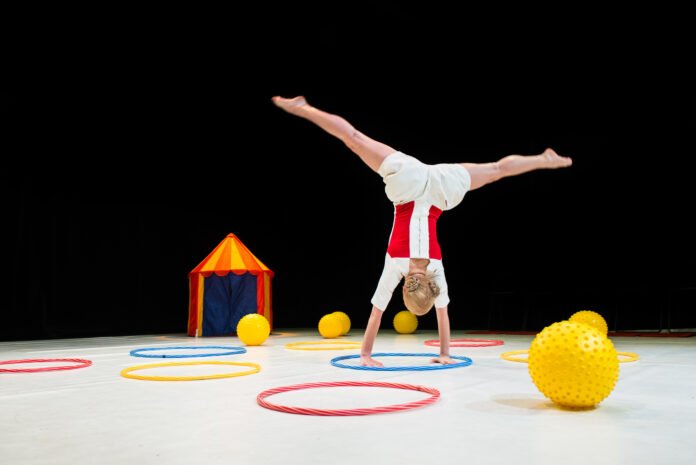 #descriçãodaimagem #pratodosverem foto da coreógrafa e educadora de dança Birute Baneviciute durante apresentação da peça teatral Colourful Games. Ela é uma mulher loira e está fazendo uma acrobacia entre bolas e bambolês.